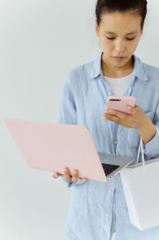 Asian woman multitasking with a pink laptop and smartphone while holding a shopping bag against a white background.
