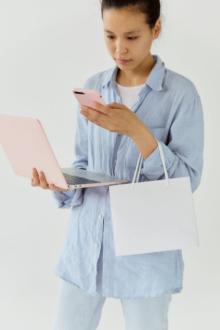 Woman Using Her Smartphone While Holding A Shopping Bag And A Laptop