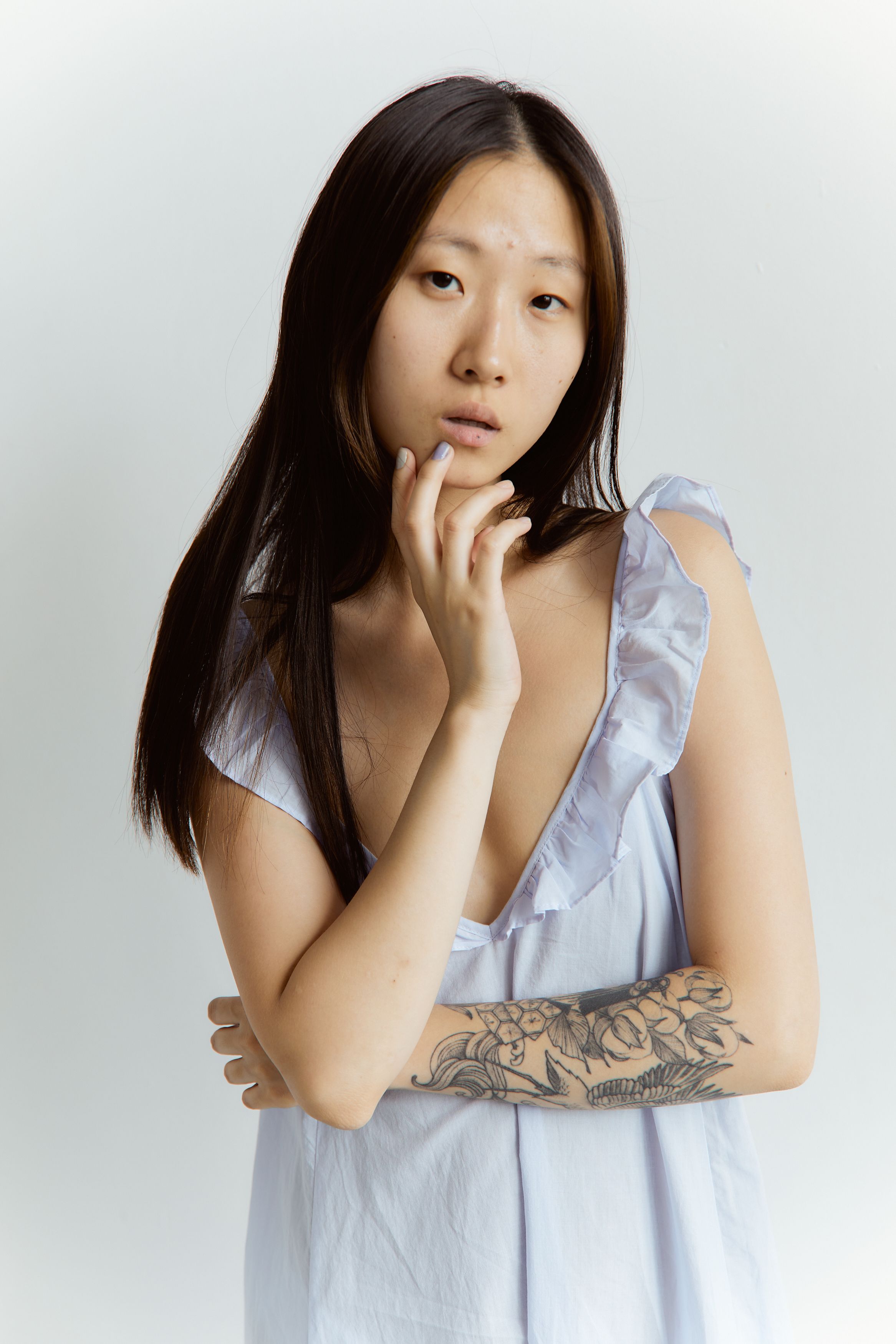 Portrait of an Asian woman with a tattoo, posing thoughtfully in a white dress against a plain background.