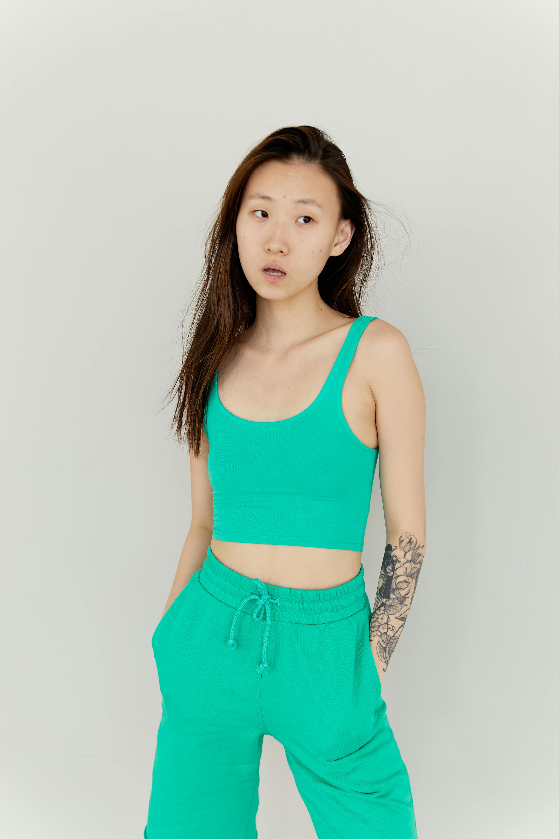 Tattooed woman in green tank top and shorts posing thoughtfully in studio setting.