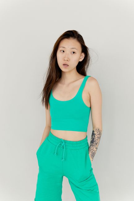 Tattooed woman in green tank top and shorts posing thoughtfully in studio setting.