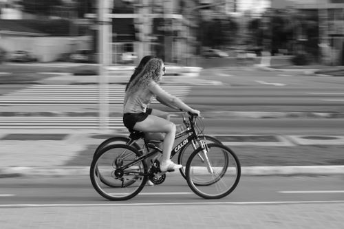 Free A young woman with curly hair rides a bicycle swiftly down a city street, captured in black and white photography. Stock Photo
