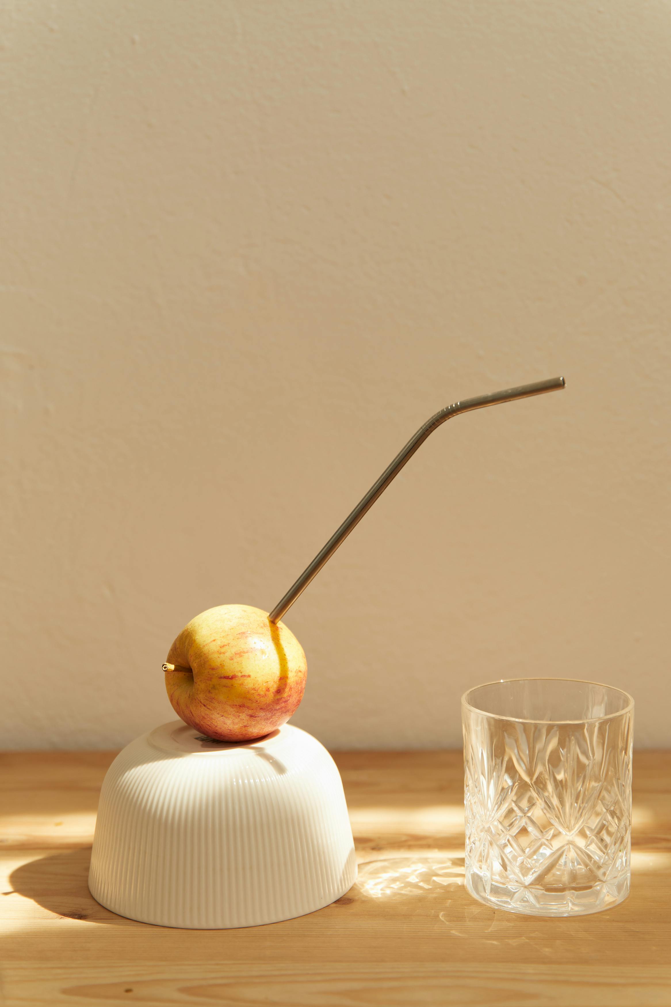 Artistic still life of an apple with a metal straw beside an empty glass.
