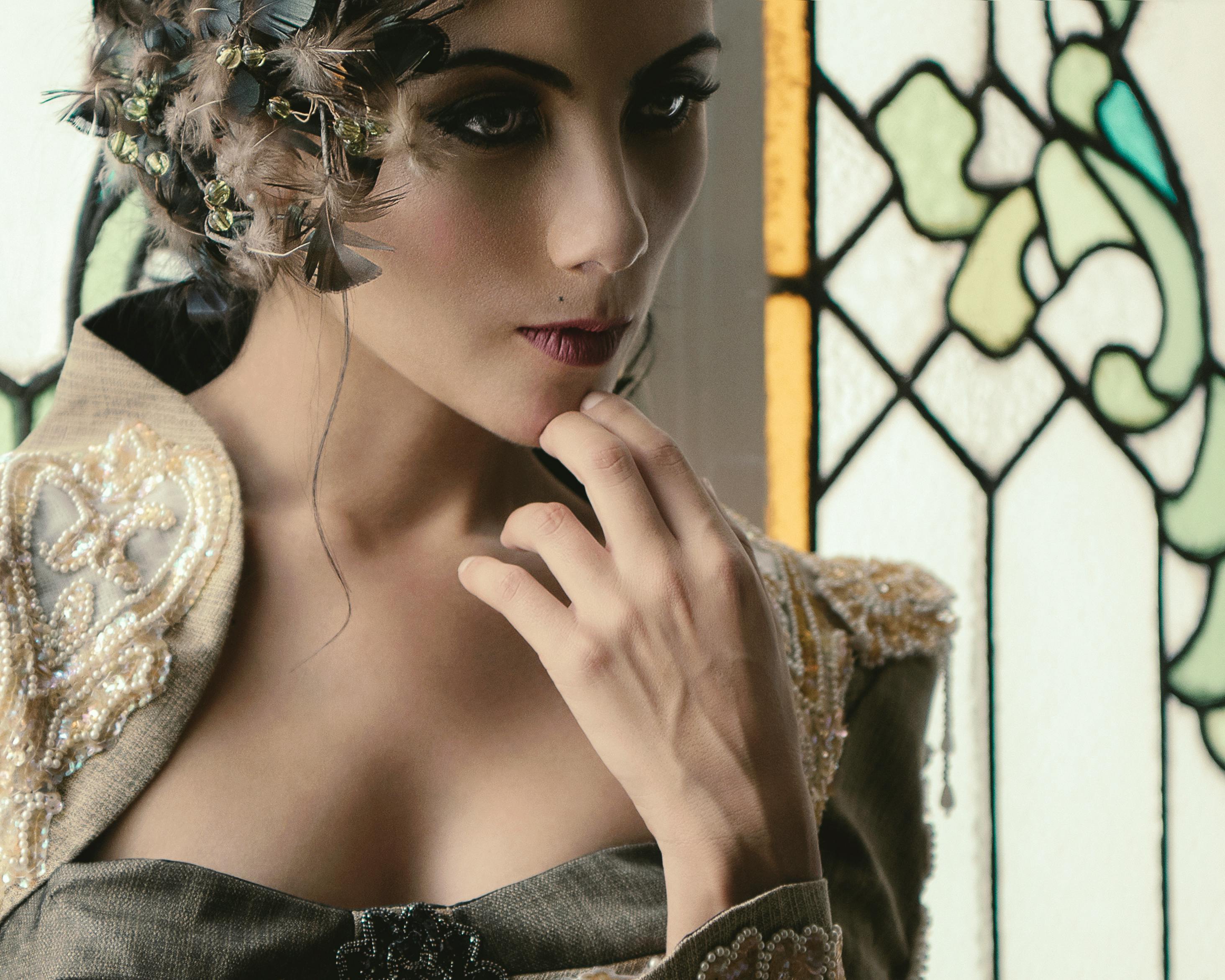 An elegant woman in artistic attire poses thoughtfully near a stained glass window.