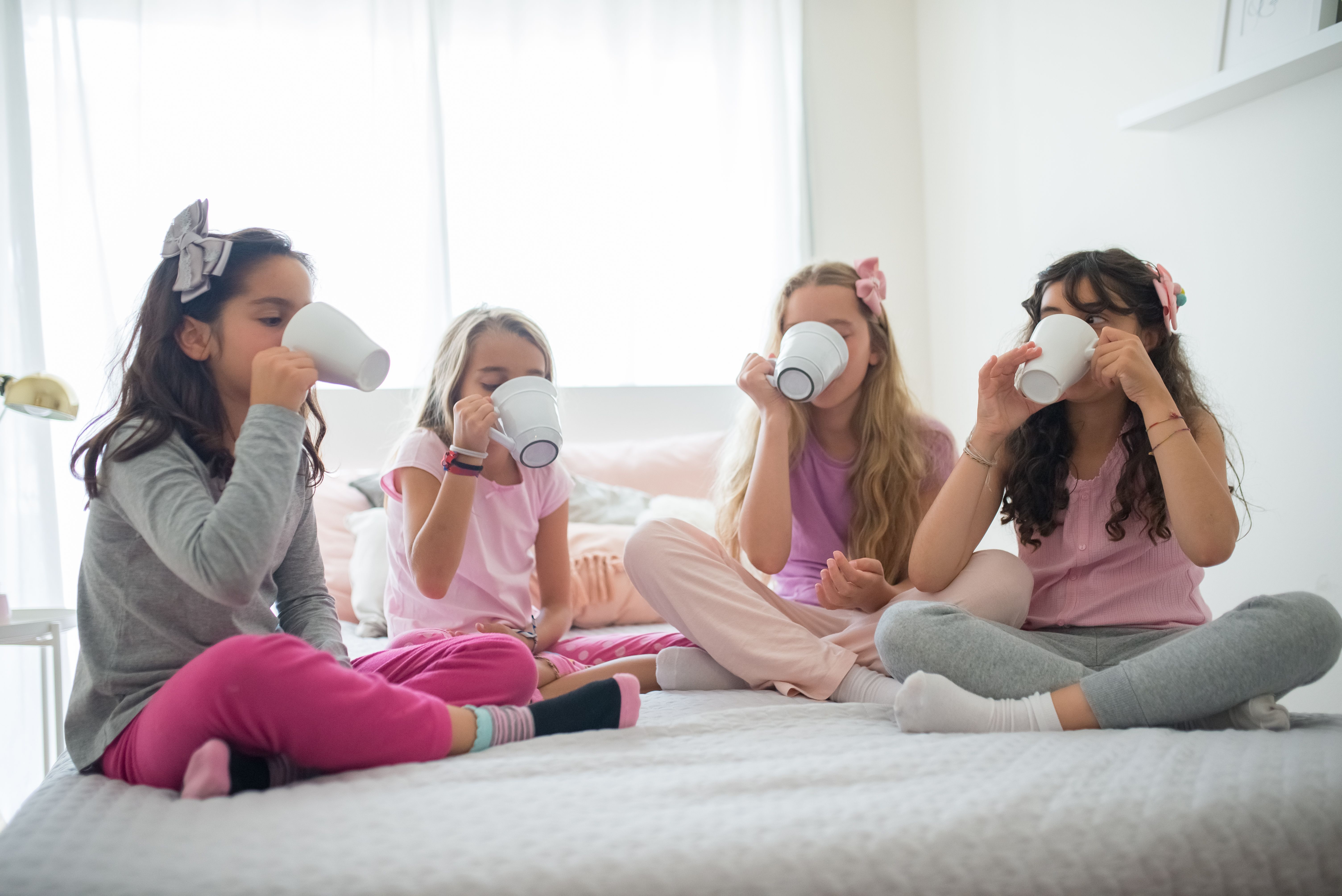 Four girls sitting on a bed, enjoying drinks together in a bright and cozy room.