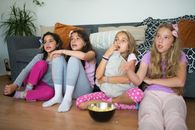 Four girls in pajamas enjoying a movie night at home, sharing popcorn on the floor.