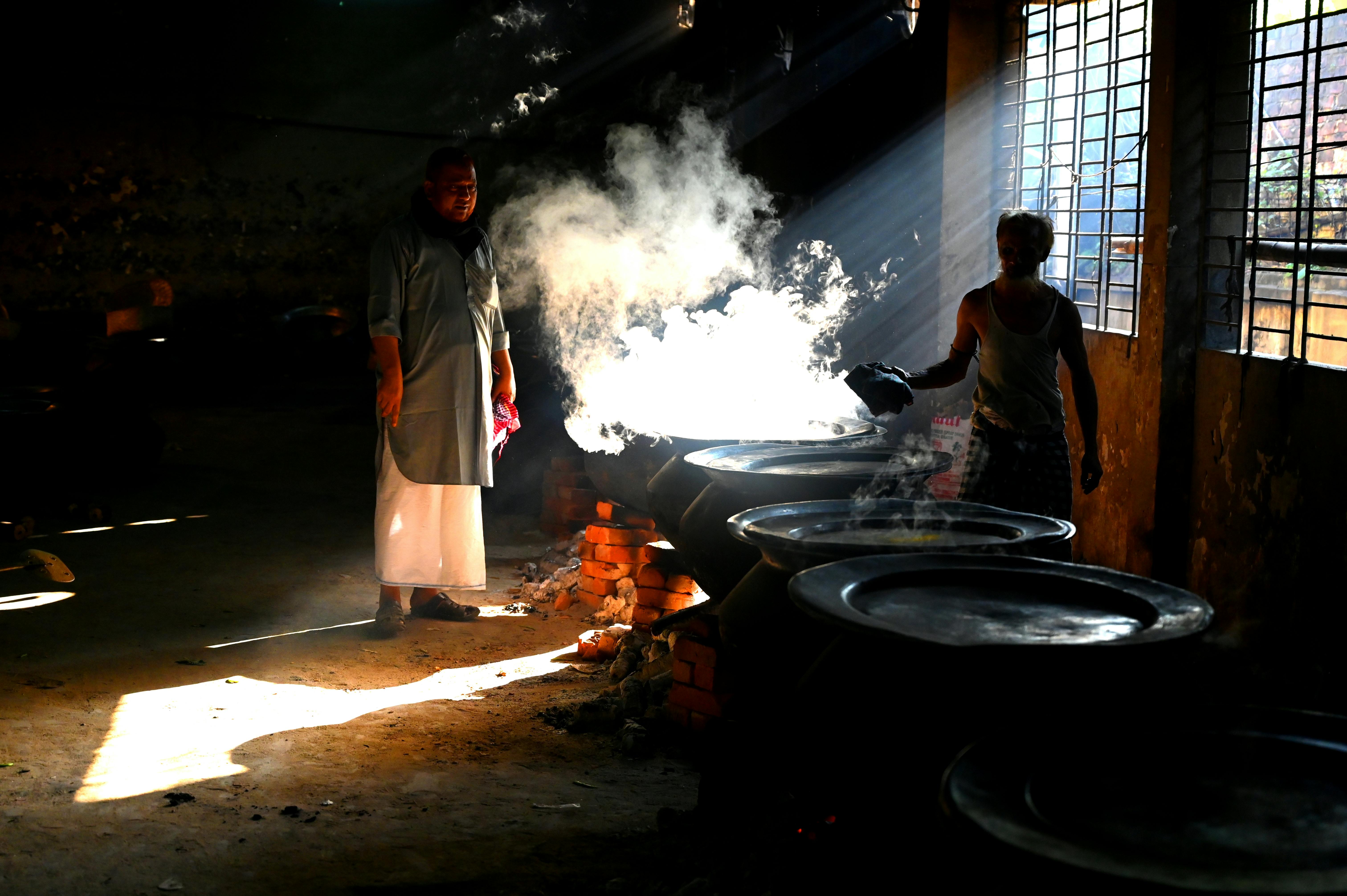 Men cooking traditional food in a steamy Chattogram kitchen with natural light.