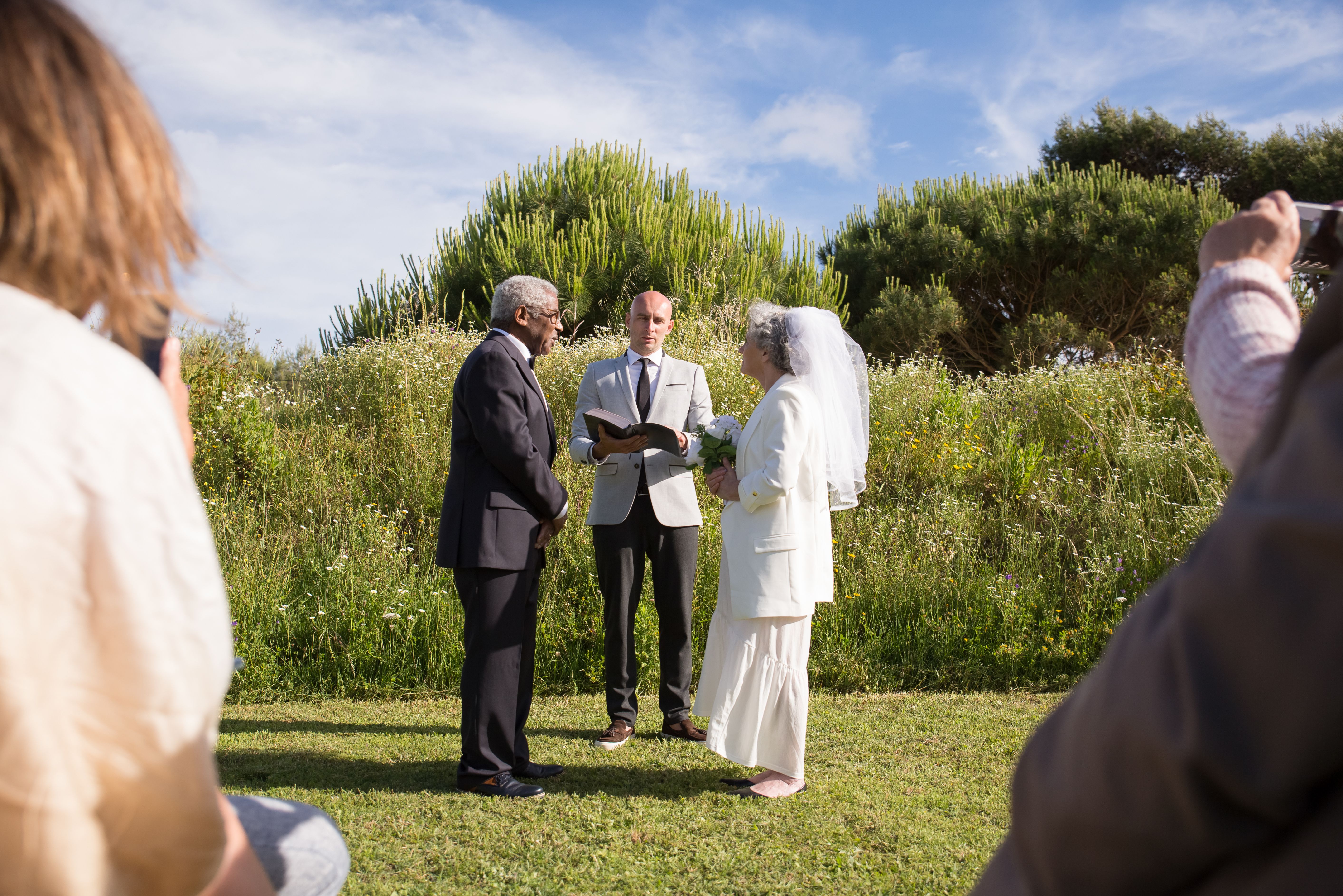 Elderly couple exchanging vows in an outdoor garden wedding ceremony in Portugal with priest officiating.
