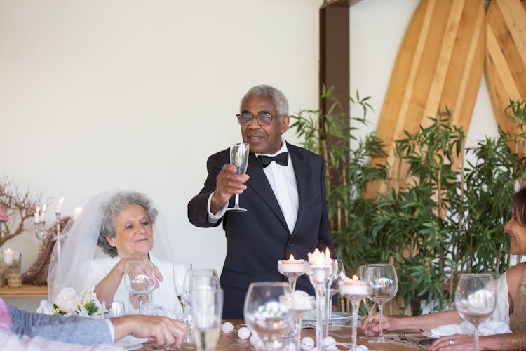 A Man In Black Suit Jacket Beside Woman In White Wedding Dress Raising His Champagne Glass