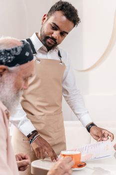 A waiter serves coffee and a menu to an elderly man seated at a cafe table.