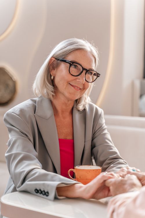 Free Senior woman in a blazer and eyeglasses holding hands in a cozy café setting. Stock Photo