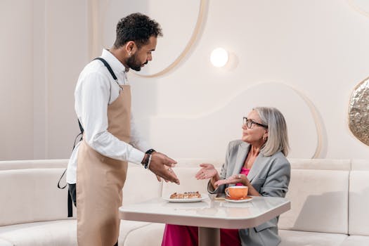 A waiter gestures while serving a senior woman in a stylish cafe setting, discussing dessert options.