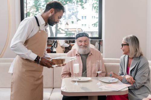 Free Elderly couple being served champagne by a waiter in a restaurant setting. Stock Photo