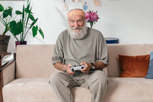 Senior man with beard playing video games on a sofa indoors, showcasing active aging.
