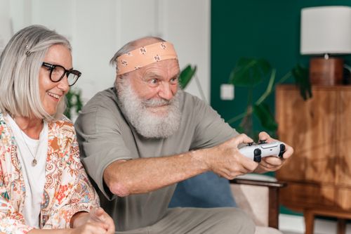Free Senior couple having fun playing video games in the living room, showcasing joy and timeless enjoyment. Stock Photo
