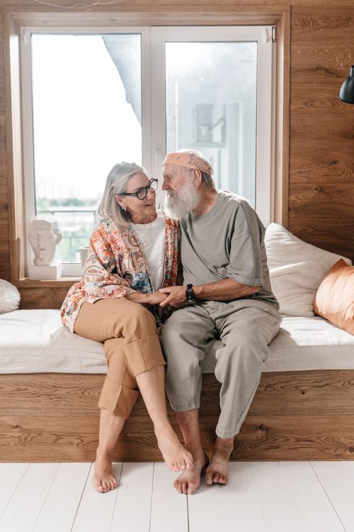 Free An elderly couple sitting together on a cozy bench in a sunlit room, expressing affection. Stock Photo