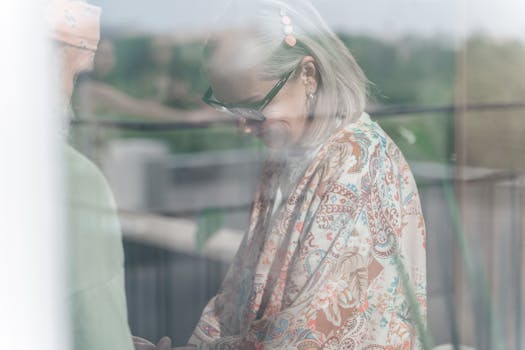 Senior woman with glasses and elegant pattern shawl smiling gently, reflected through a window.
