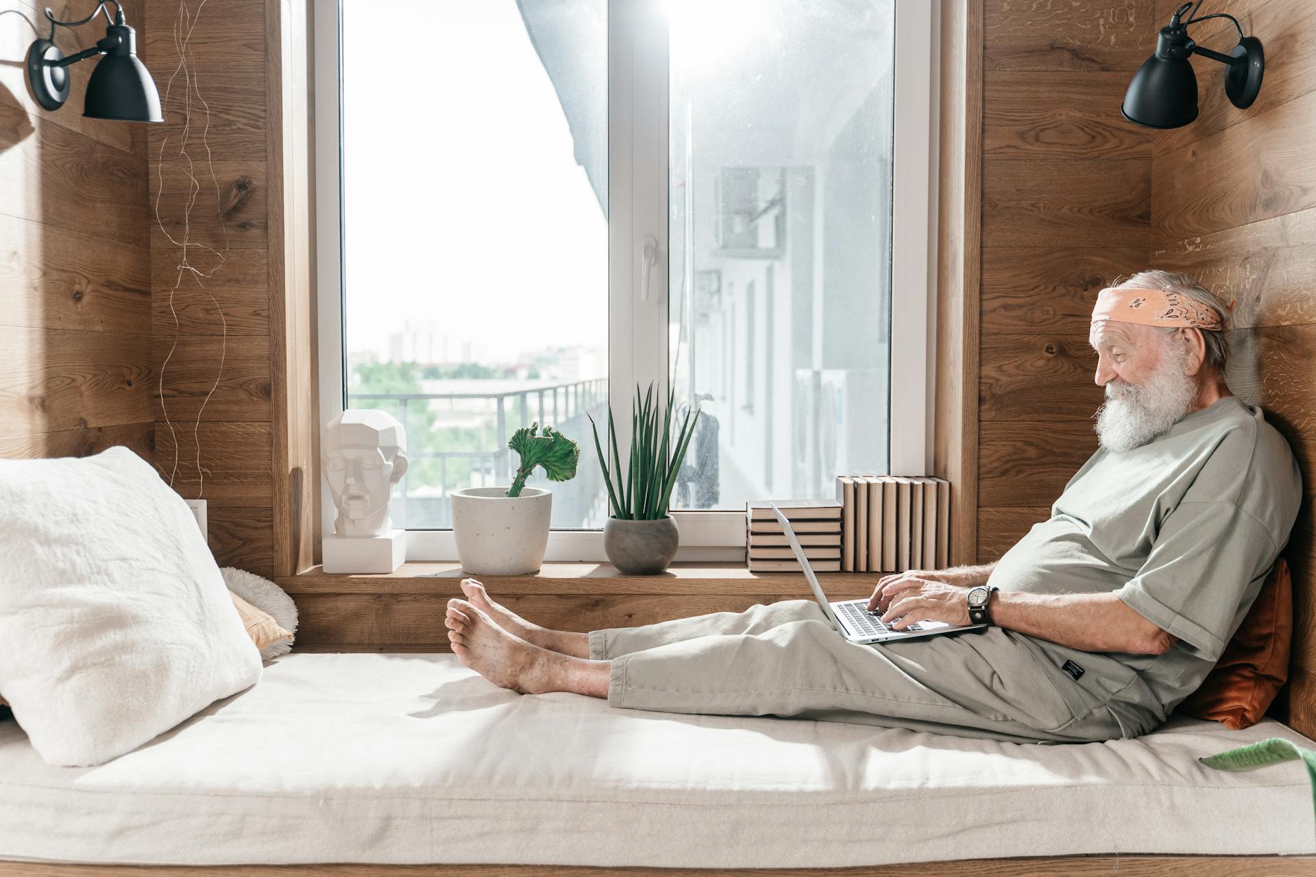 Senior man sitting comfortably at home, using a laptop by the window.