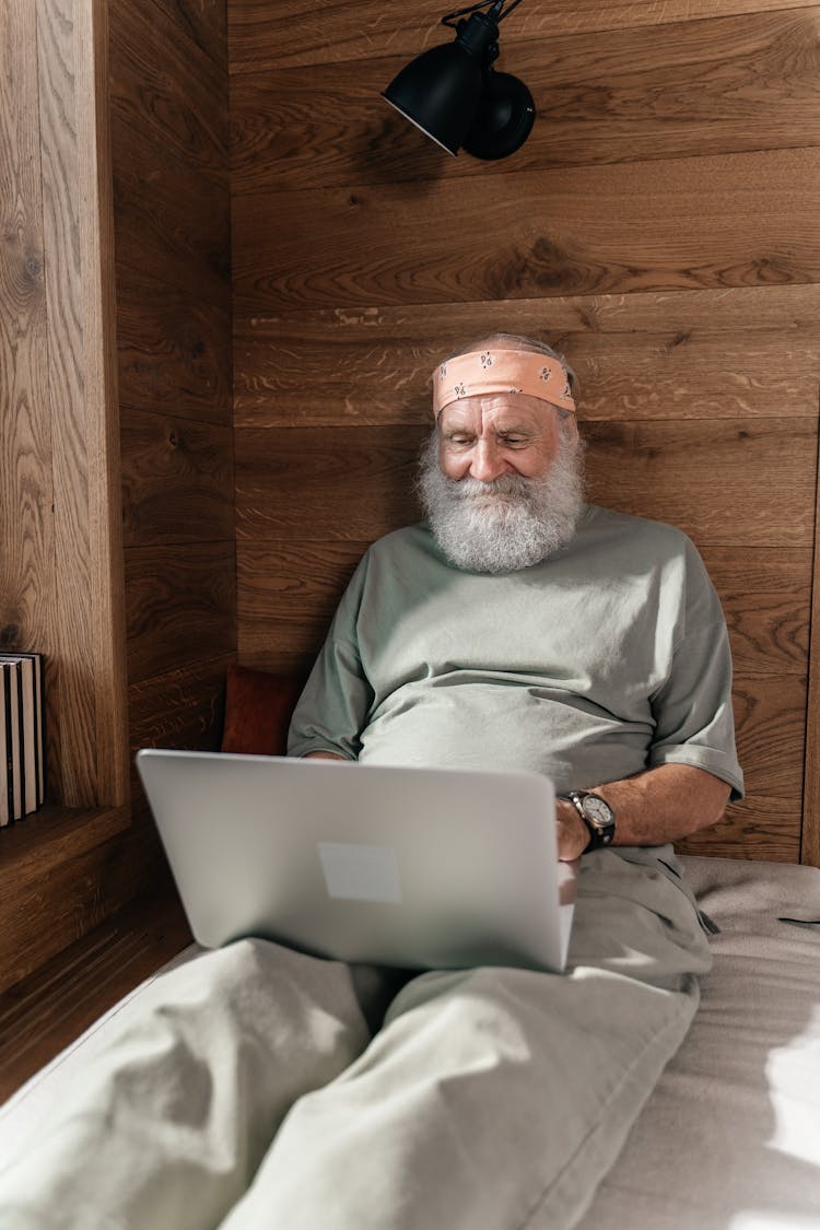 Happy Elderly Man Sitting On Bed Typing On Laptop