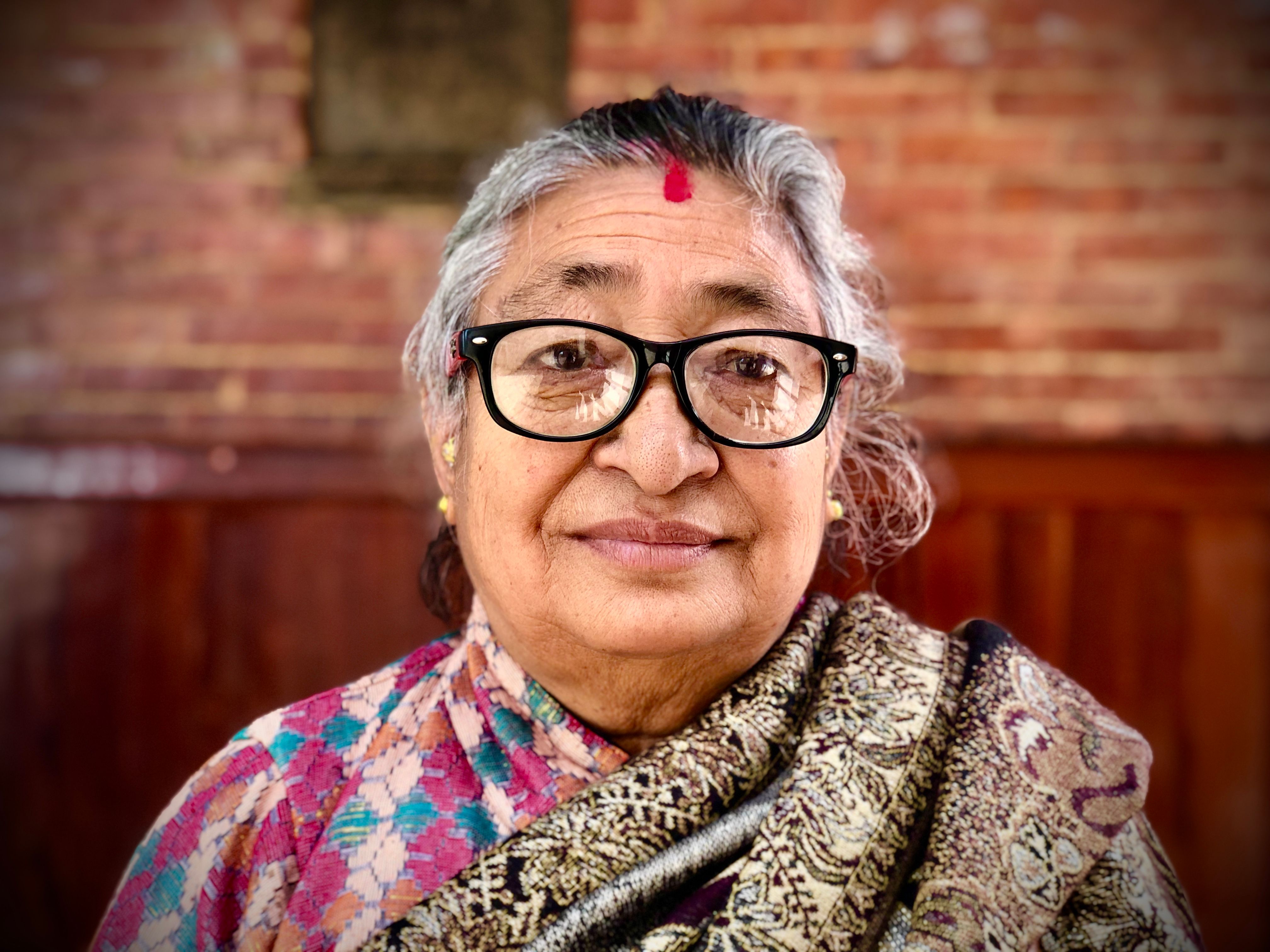 A senior woman with a vibrant Tika and eyeglasses, adorned in traditional attire, smiling warmly.