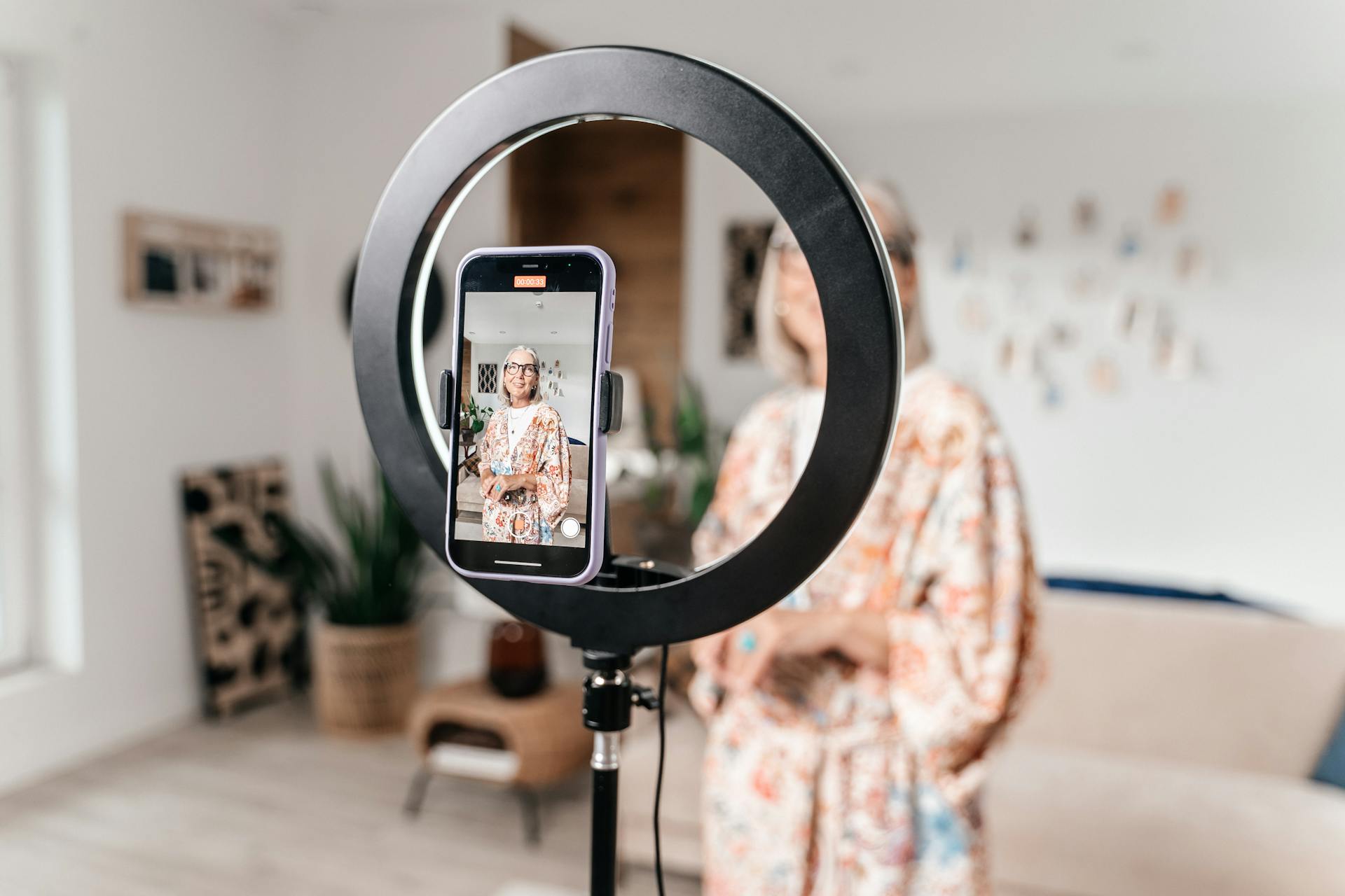 Senior woman creating content at home using smartphone and ring light in a modern setting.