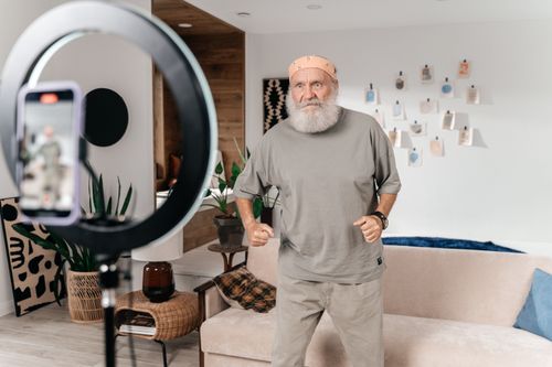 Free Senior man dances for a social media video using a phone and ring light setup indoors. Stock Photo