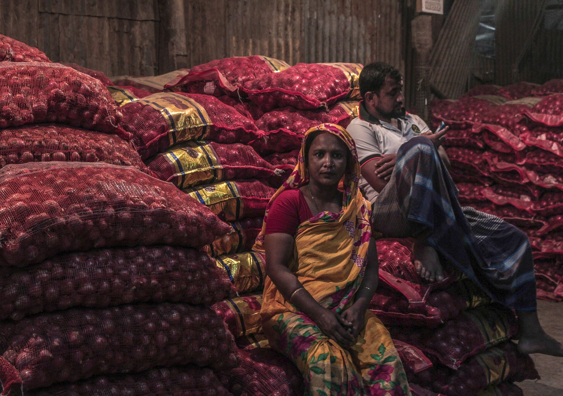 A man and woman sit among sacks of onions in a market in Dhaka, Bangladesh, captured in a candid moment.