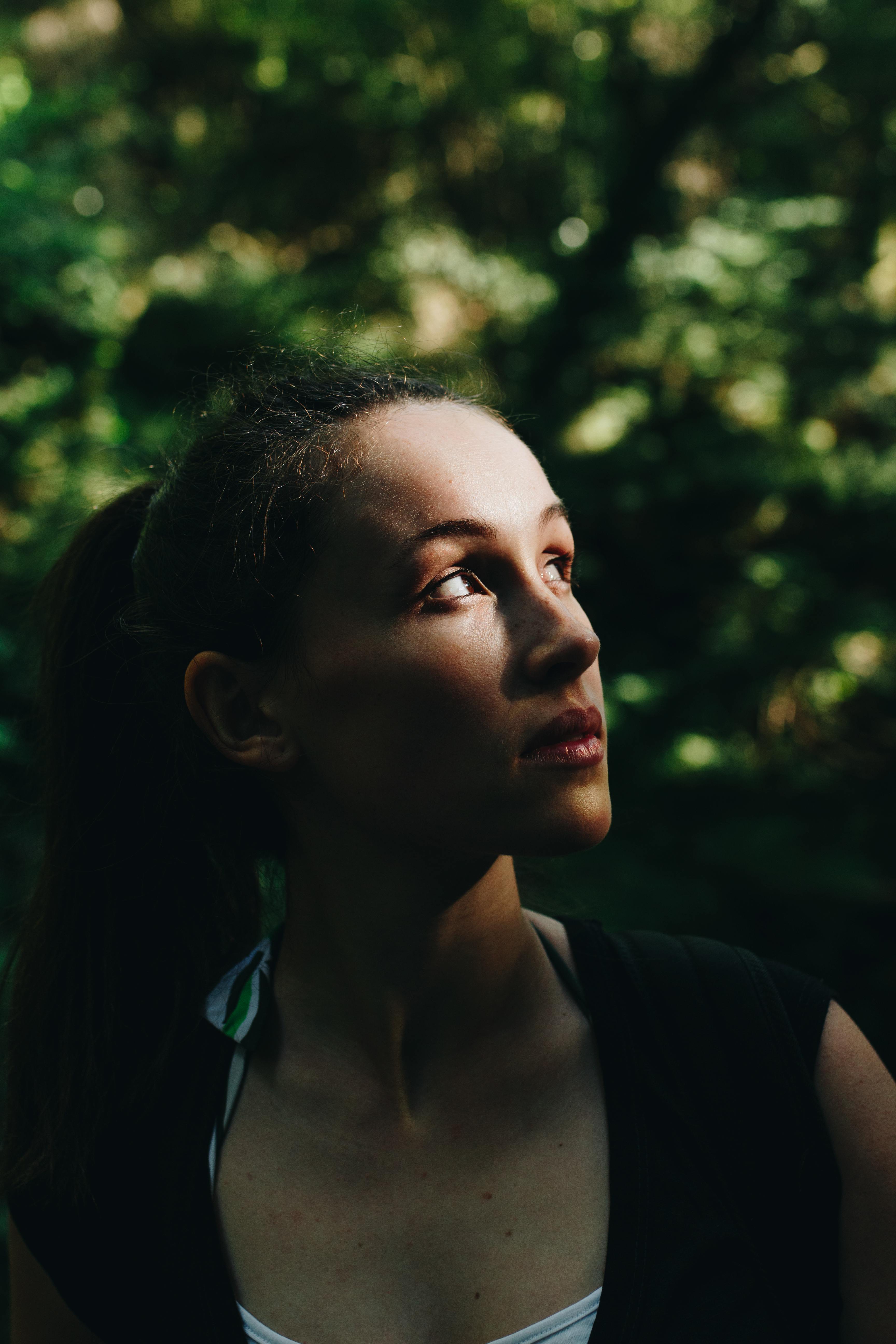 A thoughtful young woman looks up in a forest with warm sunlight casting on her face.