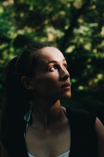 A thoughtful young woman looks up in a forest with warm sunlight casting on her face.