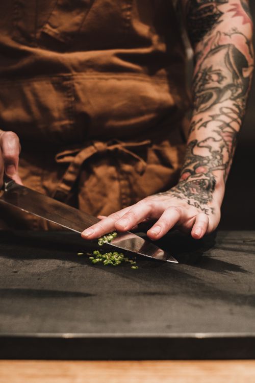 Free A tattooed chef's hands skillfully chopping herbs on a dark cutting board, highlighting culinary artistry. Stock Photo