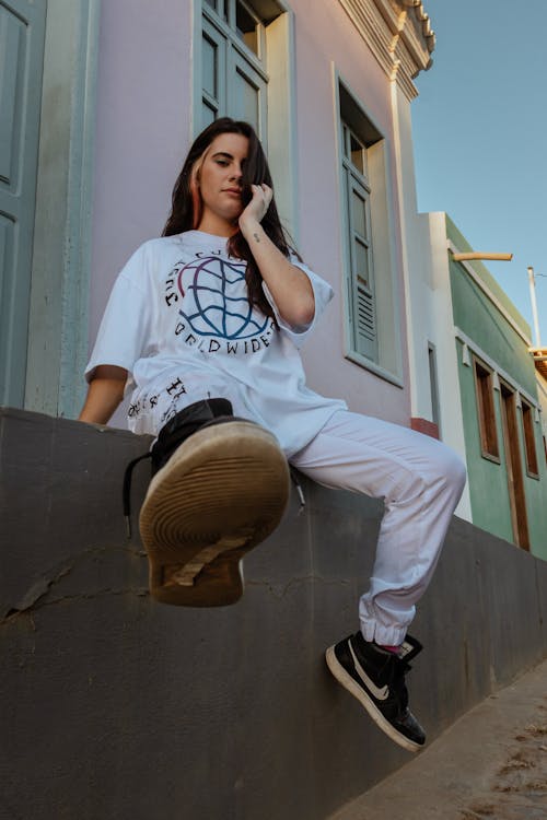 Woman in White Crew Neck T-shirt and White Pants Sitting on Concrete Wall 