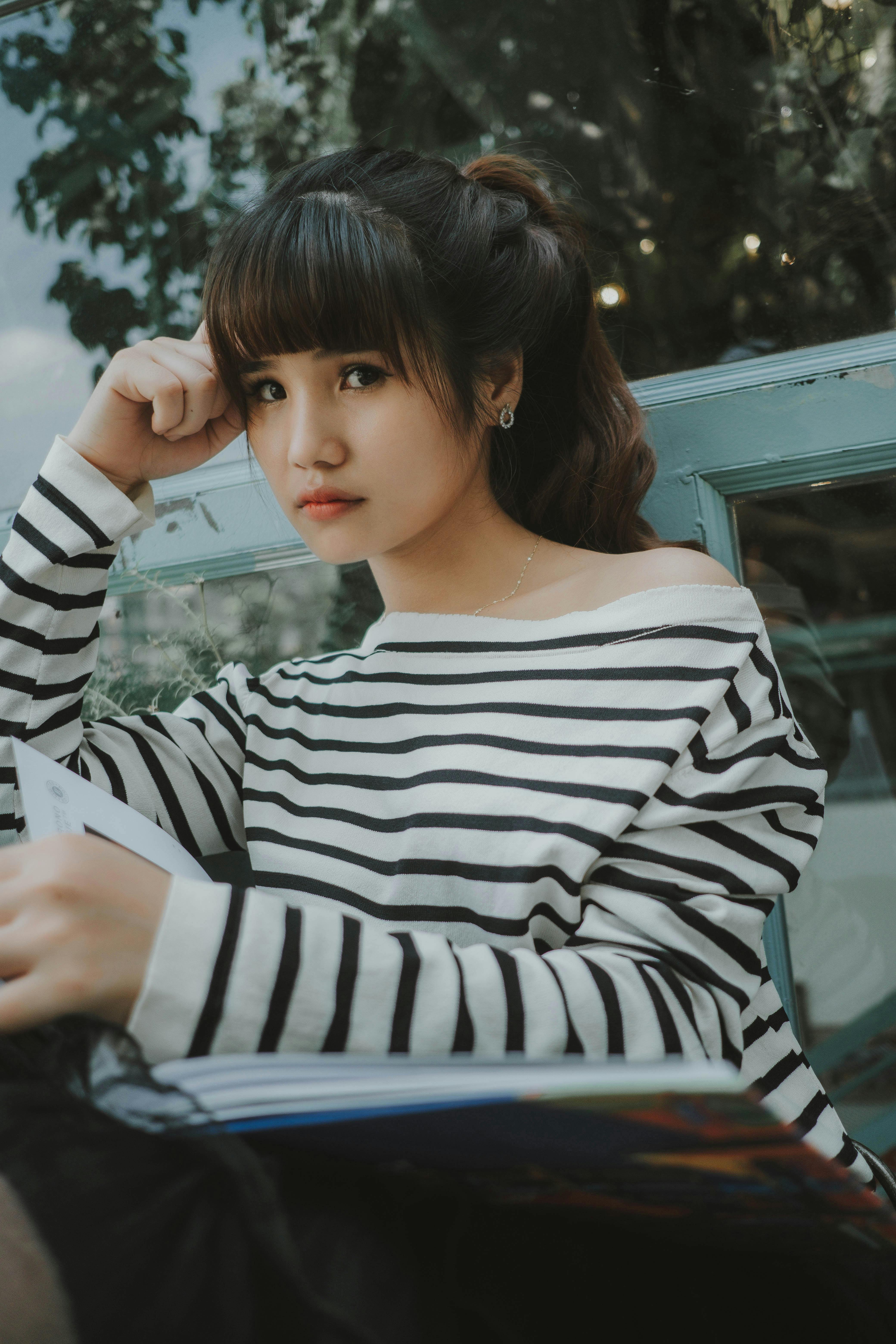 Asian woman in a striped shirt sitting outdoors with a book, exuding a relaxed and stylish vibe.