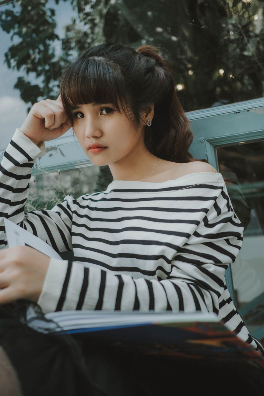 Asian woman in a striped shirt sitting outdoors with a book, exuding a relaxed and stylish vibe.