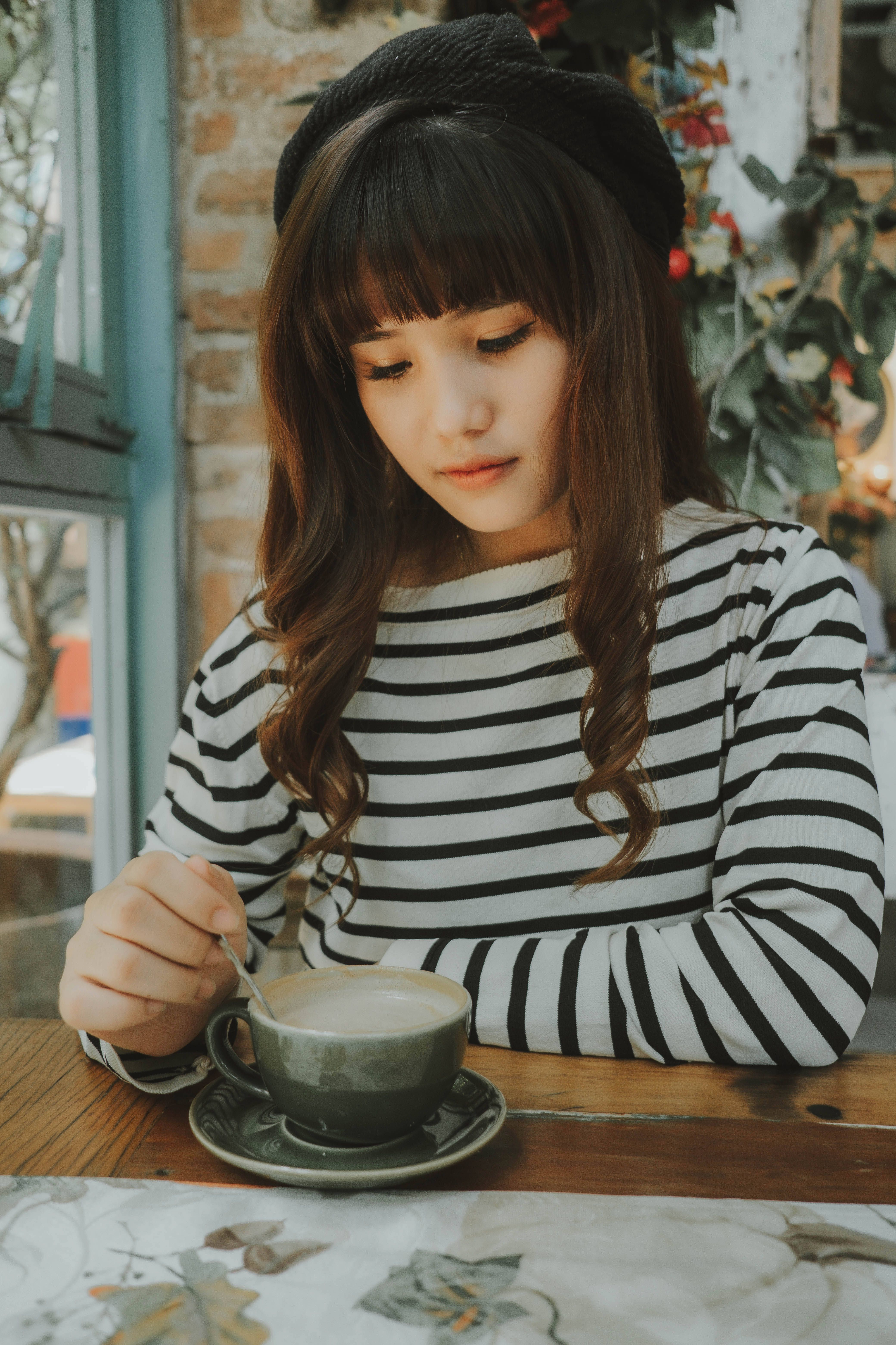 Stylish young woman in striped shirt sipping coffee at a cozy cafe, enjoying a quiet moment indoors.