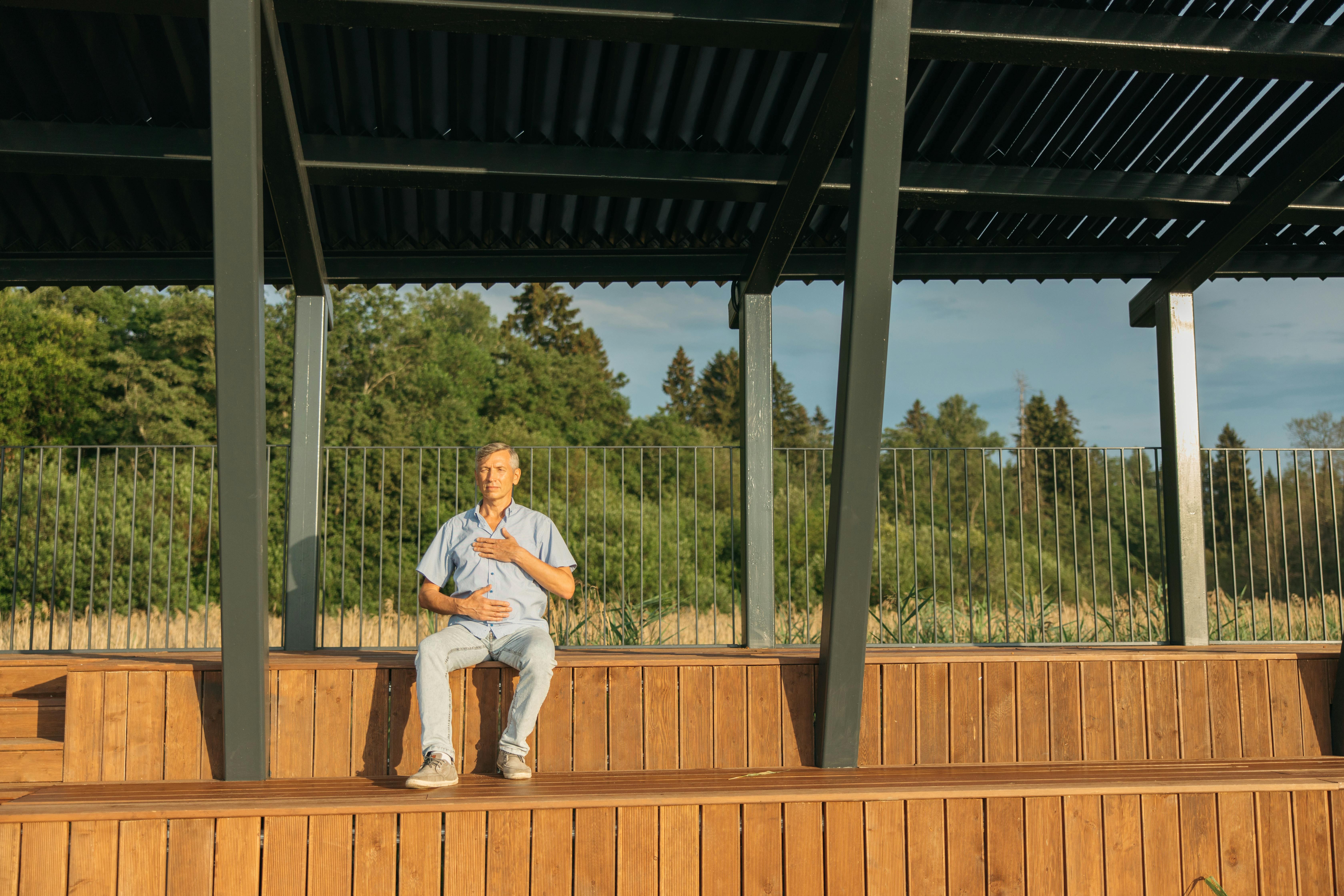 Middle-aged man practicing meditation outdoors on wooden bleachers under a metal structure in sunny weather.