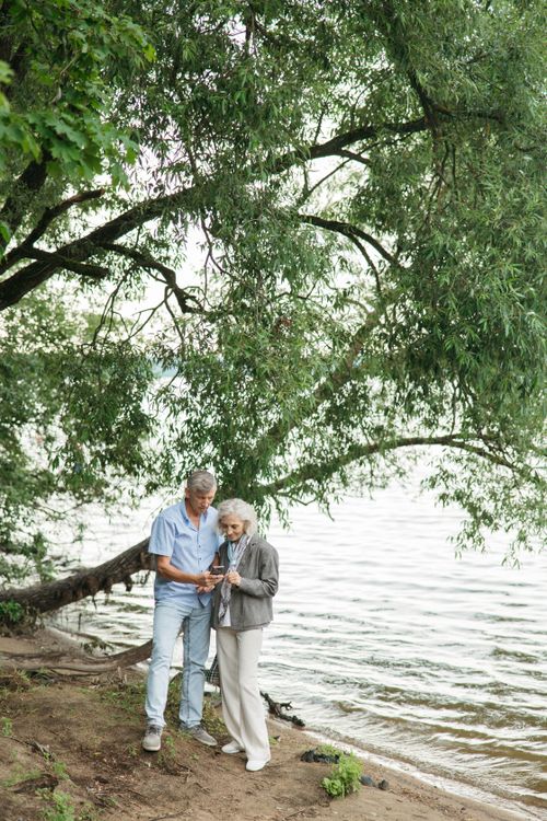 Free An affectionate elderly couple standing by a serene lakeside, enjoying a quiet moment under lush greenery. Stock Photo