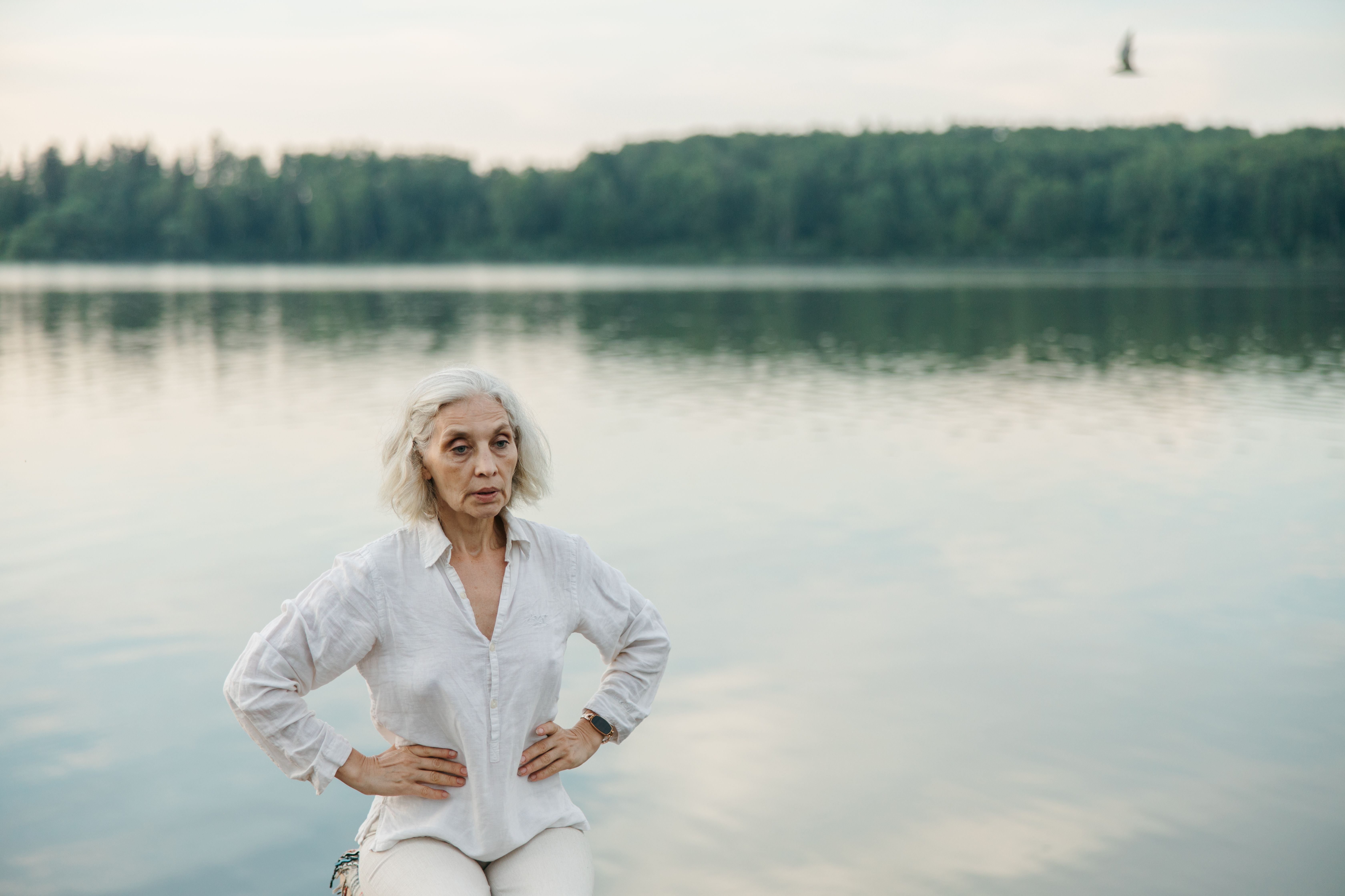 Elderly woman practicing mindfulness by a serene lake amid nature\'s tranquility.