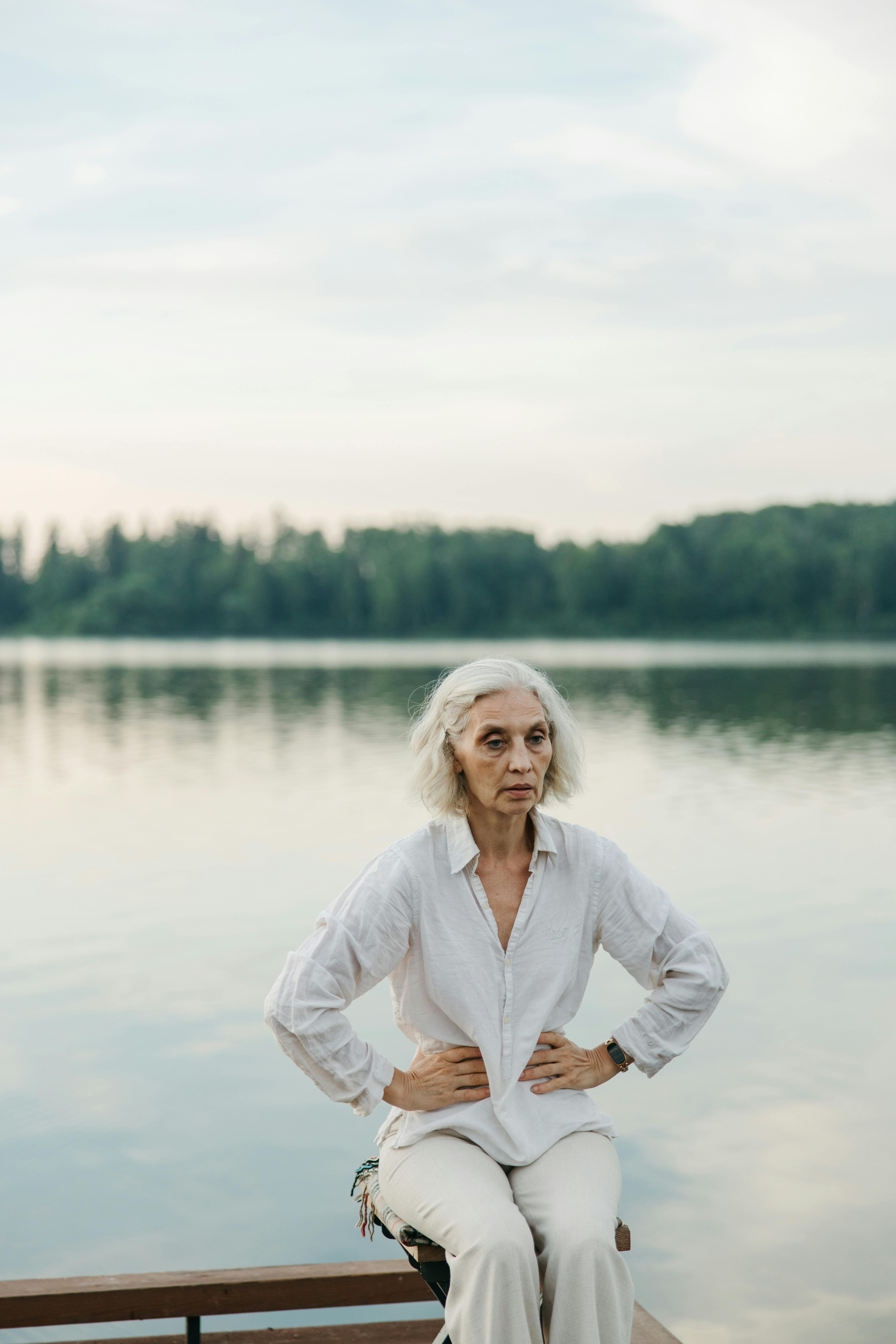 An elderly woman relaxes by a tranquil lake, embracing peaceful surroundings.
