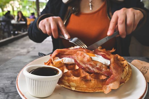 Close-up of a waffle with bacon, served with syrup in an outdoor cafe setting.