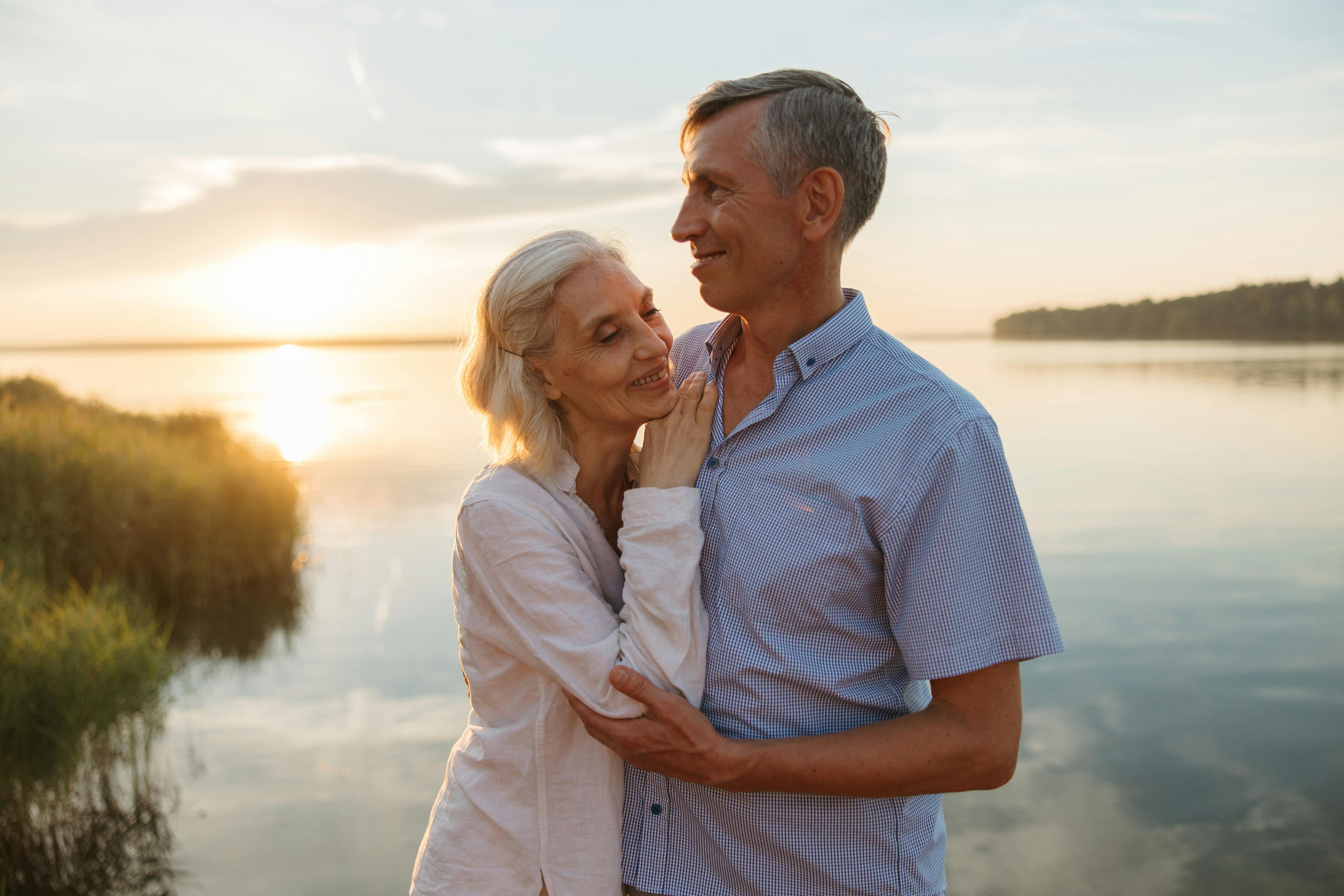 Couple embracing by a lake at sunset. The woman rests her head on the man's shoulder.