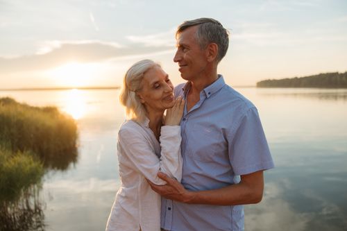 Free A senior couple lovingly embraces by a serene lake during a beautiful sunset. Stock Photo