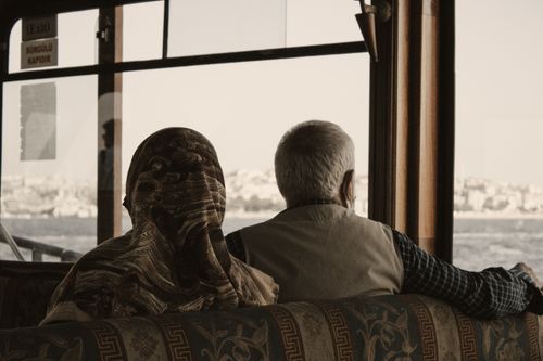 Free Elderly couple sitting side by side on a ferry, looking outside at a scenic cityscape through large windows. Stock Photo