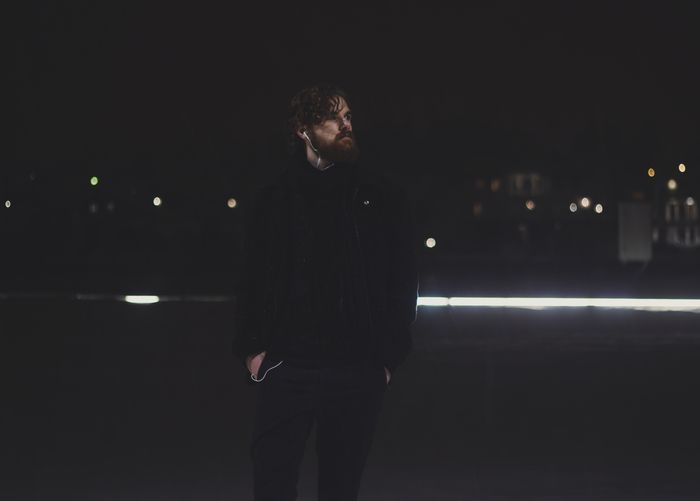 A man with facial hair stands stylishly outdoors at night, illuminated by dim lights., by Sindre Fs on Pexels