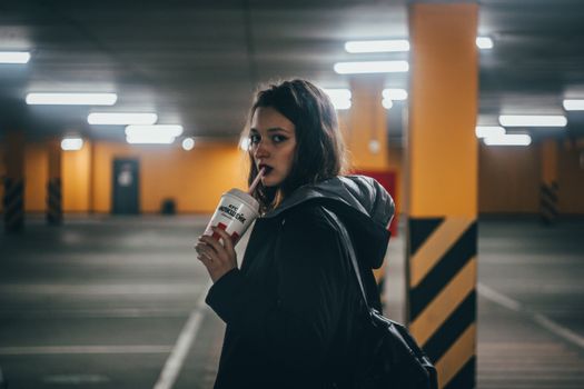 Young woman drinking a soft drink in a dimly lit parking area, adding a cool and moody vibe.