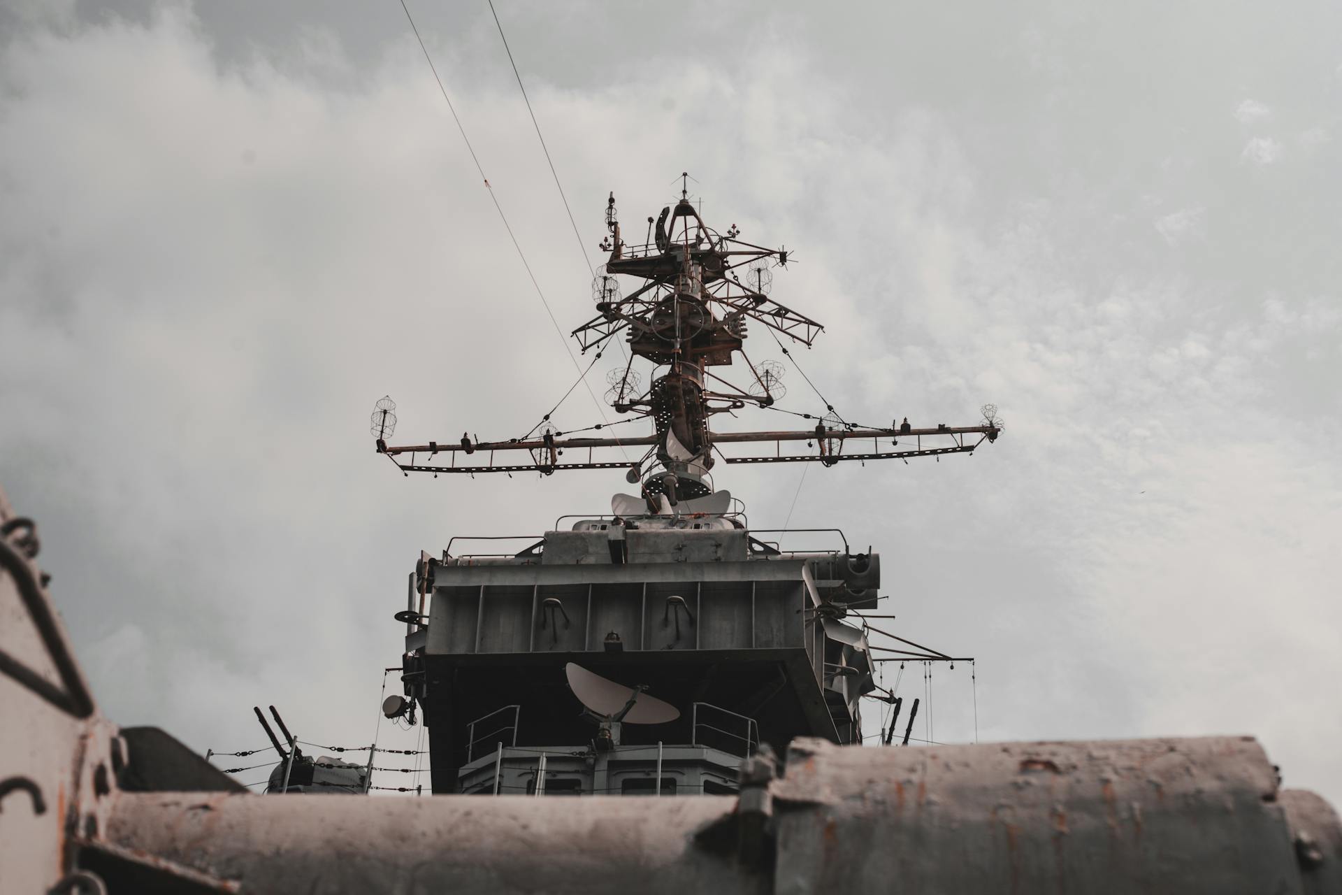 Close-up view of a military warship's radar and antenna mast against a cloudy sky.