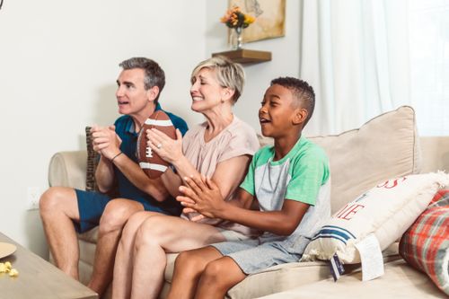 Free A joyful family watches a football game on the sofa, cheering with a football in hand. Stock Photo