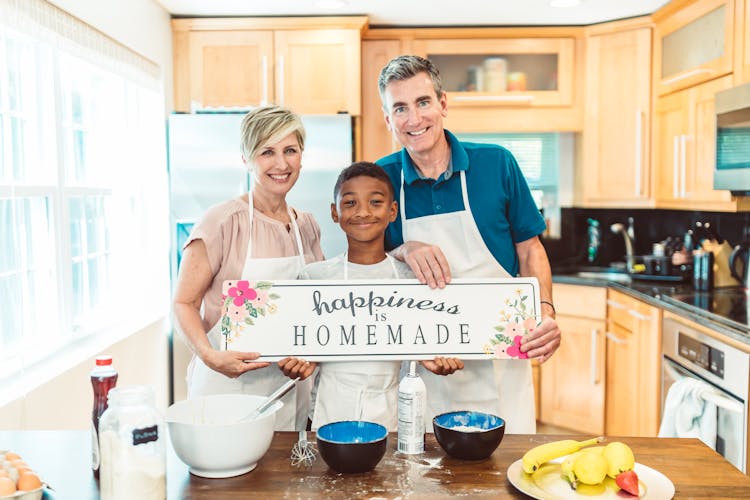 A Happy Family Wearing Apron Standing In The Kitchen While Holding A Signage