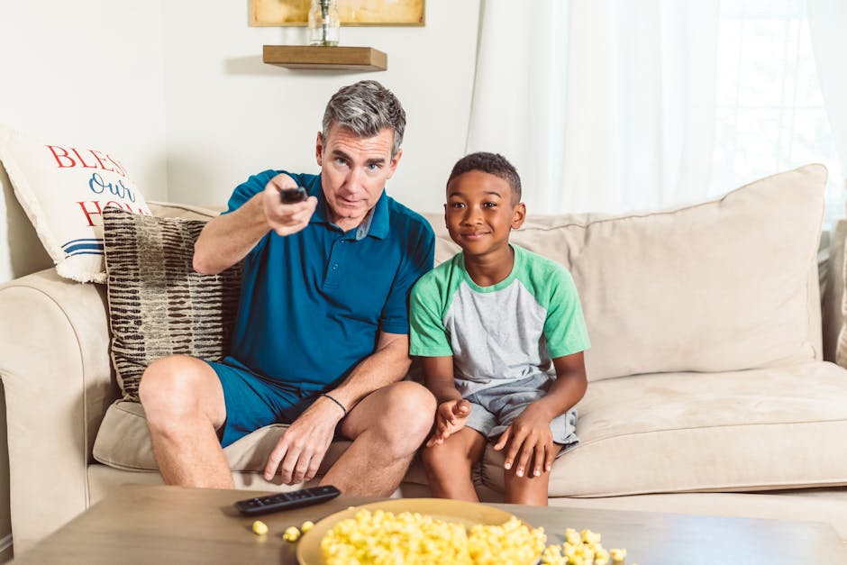 Father and son sitting on a sofa enjoying a movie at home with popcorn.
