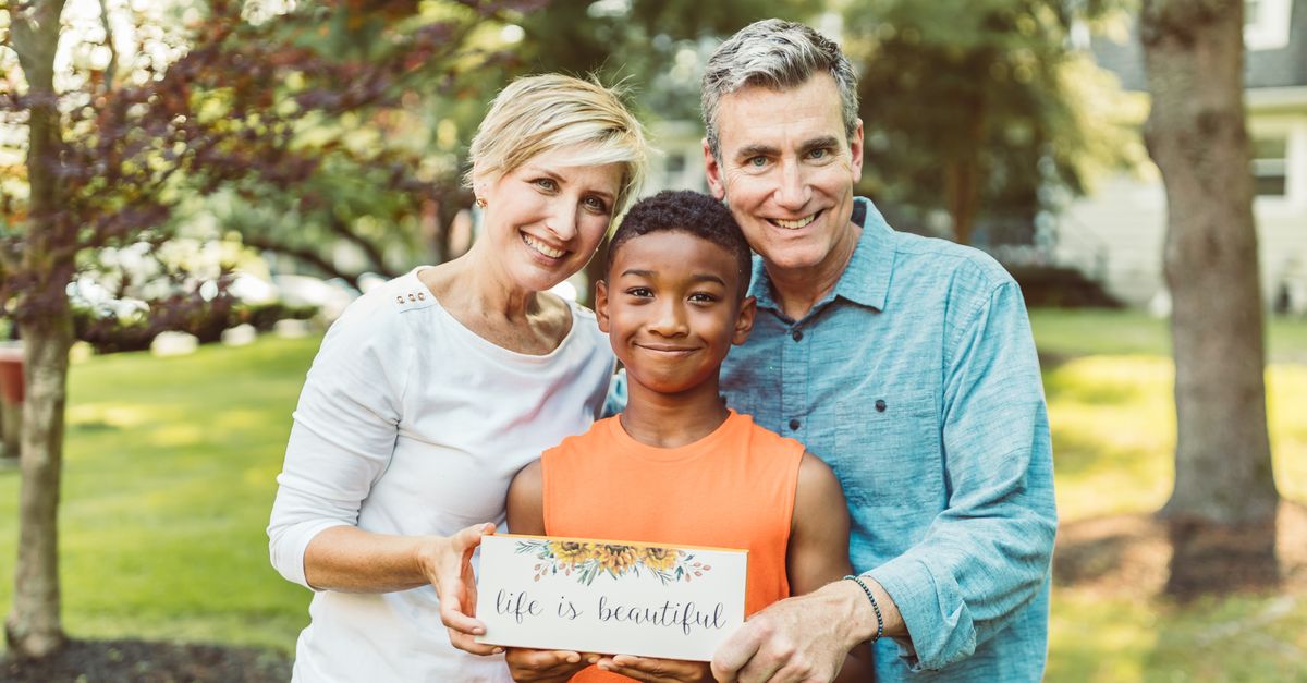 A joyful family portrait with parents and their adopted child holding a 'life is beautiful' sign.