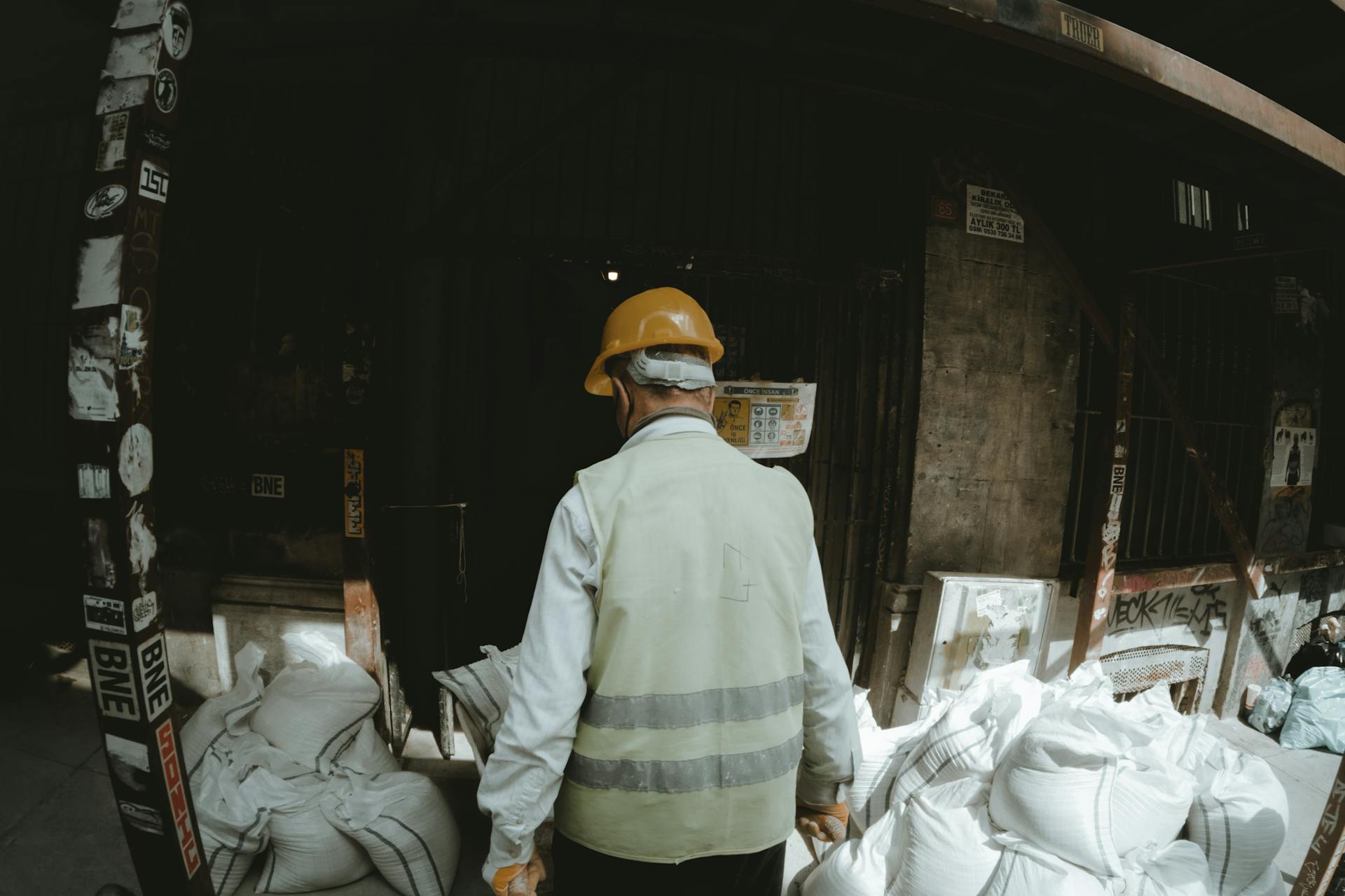 Construction worker wearing hard hat and vest surrounded by white sacks in an urban environment.